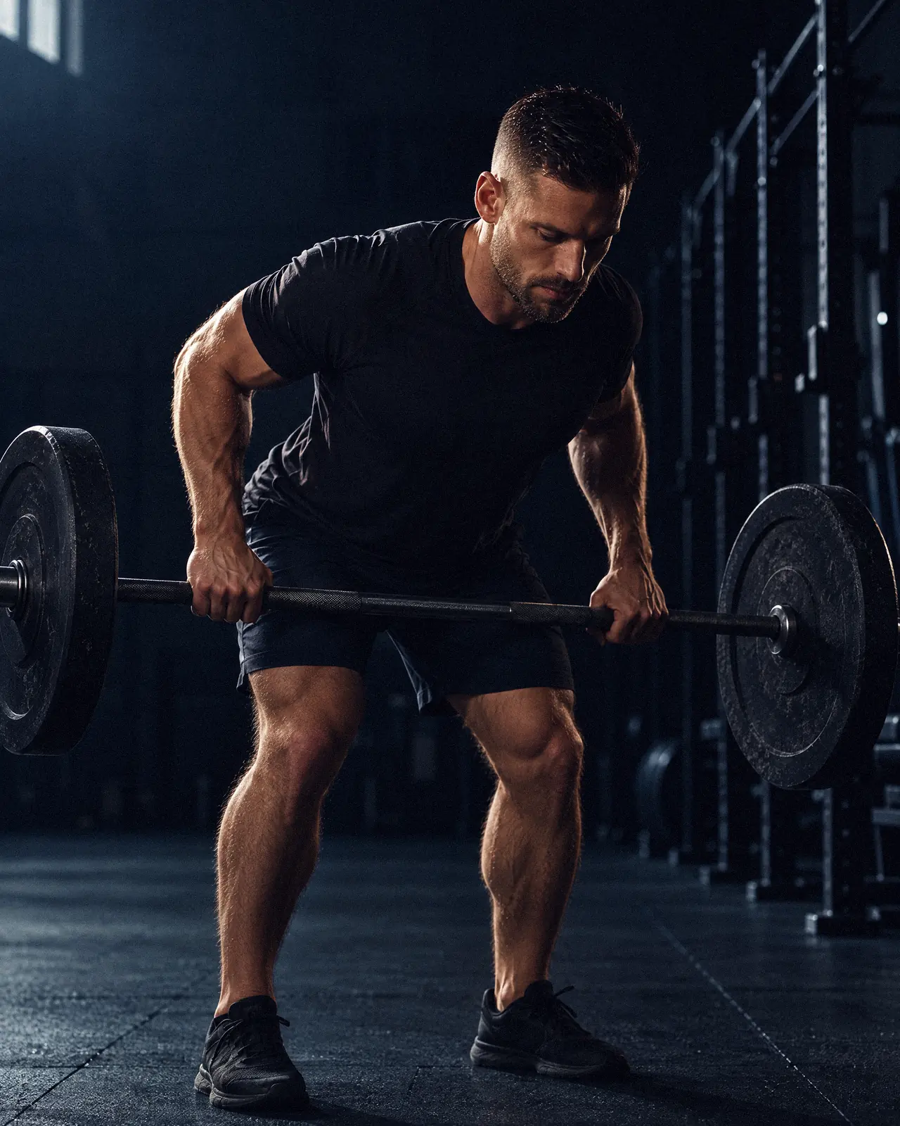 A man mid-rep of a bent-over barbell row in a dark gym, lit from behind.